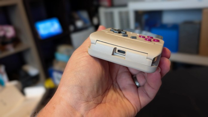A person holding a small, portable gaming device with buttons and a USB-C port, set against a blurred background of a workspace.