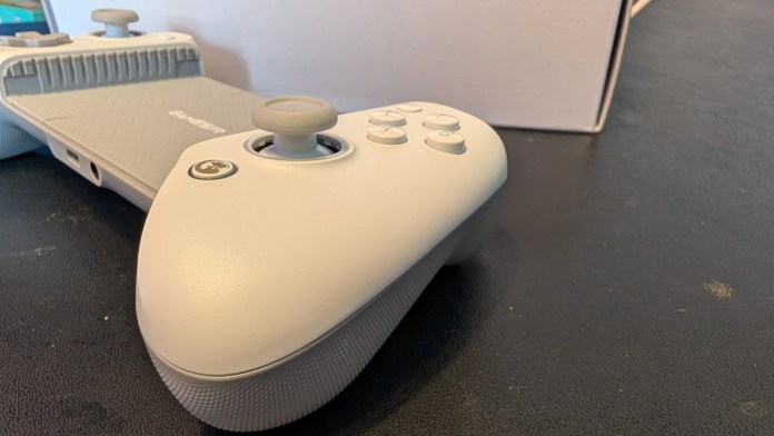 Close-up view of a gray gaming controller featuring a joystick, buttons, and textured grip, placed on a black surface with a partially visible white box in the background.