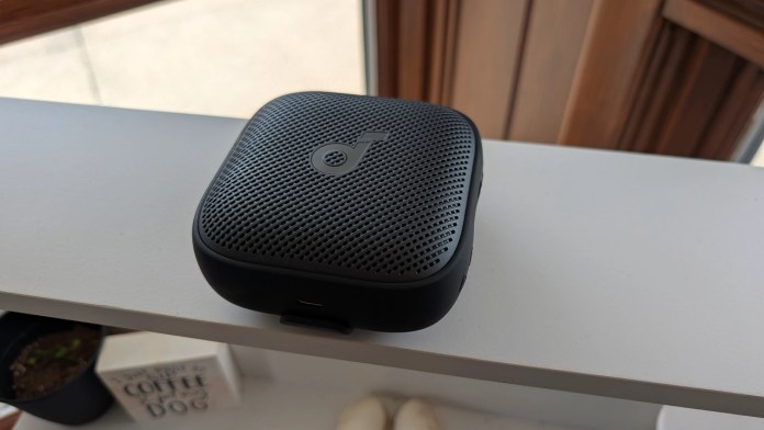 A black portable speaker resting on a white shelf, with a textured surface and a small port visible on the side.