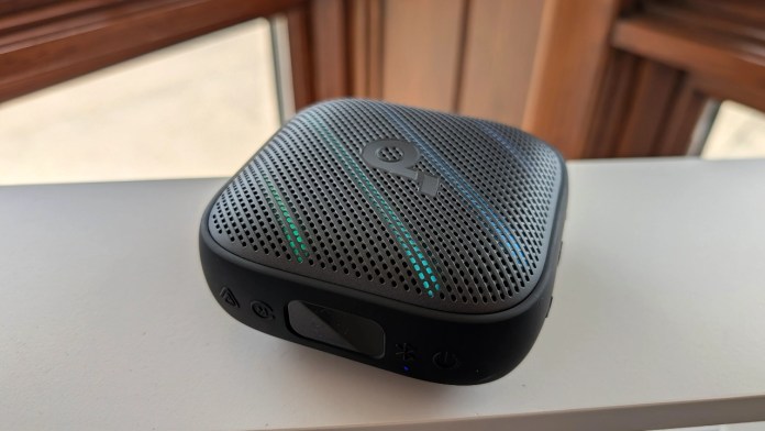 A compact wireless speaker with a perforated design and LED lights, placed on a white surface near a window.