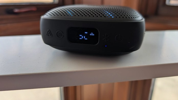 A close-up view of a portable Bluetooth speaker with a digital display showing the volume level and bass settings, placed on a white shelf.