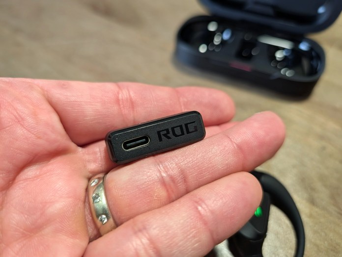 A person holding a small black USB-C connector with the 'ROG' logo, while a pair of wireless earbuds and their case are blurred in the background.