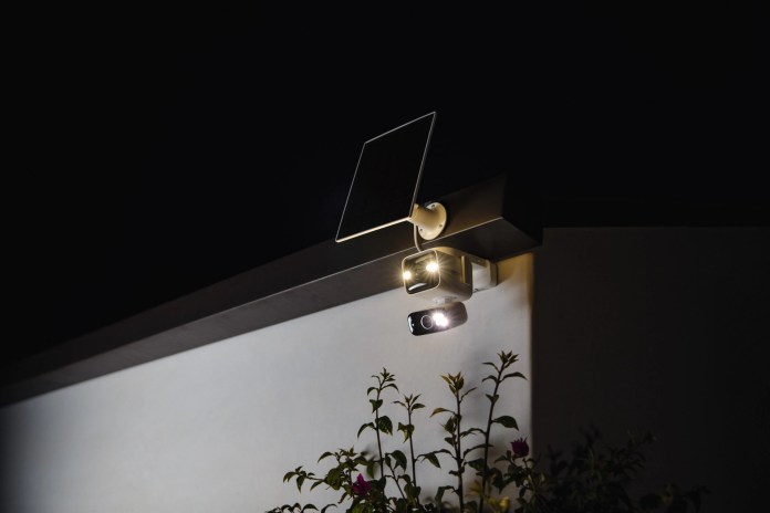 A solar-powered security light mounted on a wall, featuring a camera and a spotlight, with plants visible in the foreground, set against a nighttime background.