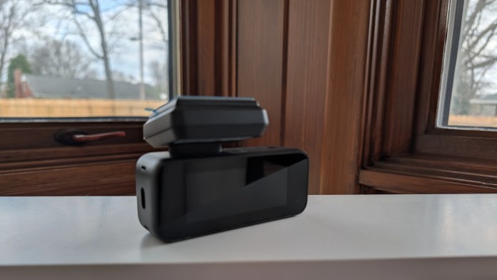 A black electronic device with a screen, placed on a white surface next to a window, showing a view of trees and a house outside.