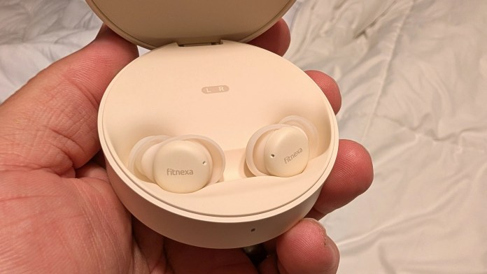 A person's hand holding a closed charging case with two white wireless earbuds inside, labeled 'fitnexa'.