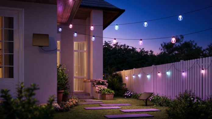 A beautifully lit backyard patio at dusk, featuring colorful string lights hanging above. The space includes a pathway of stone slabs, potted plants, and flower beds, with a white fence in the background.
