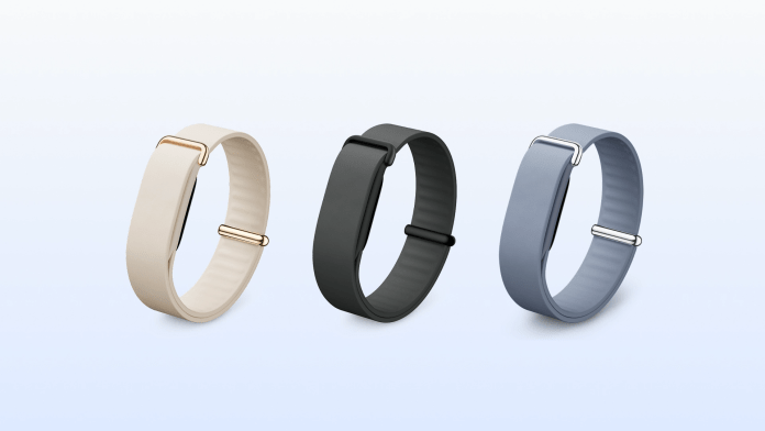 Three fitness trackers displayed in a row: one beige with gold clasp, one black, and one light blue with silver clasp, set against a soft gradient background.