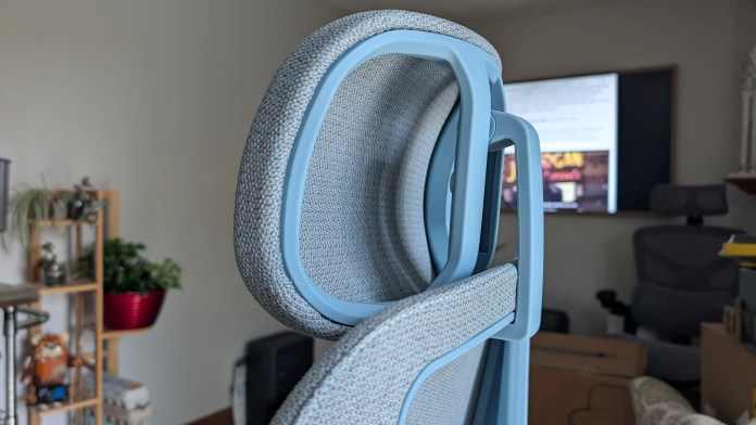 Close-up view of a modern office chair with a gray fabric seat and light blue frame, set against a background of a home office with plants and a television.