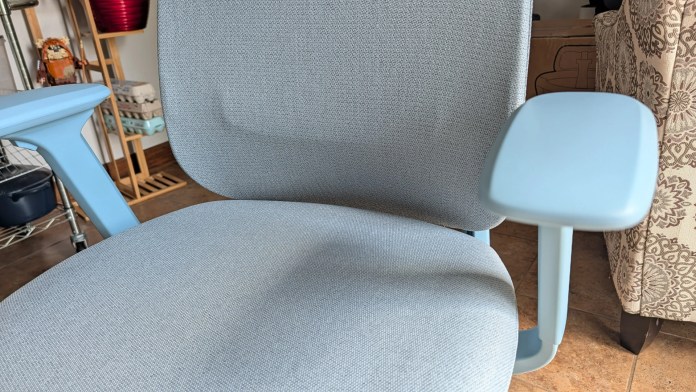 Close-up view of a modern ergonomic chair with a light blue fabric seat and armrests, placed in a cozy room setting.