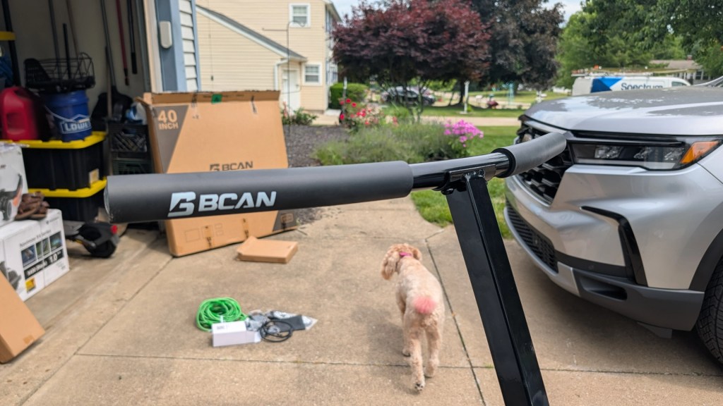 Close-up view of a BCAN handlebar attached to a black scooter, with a light-colored dog walking in the background, and a gray vehicle parked nearby.