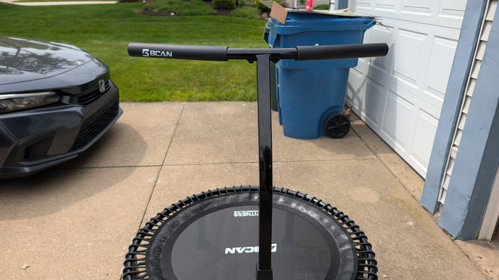 A black rebounder trampoline with safety handles in front of a parked gray car, situated on a concrete driveway.