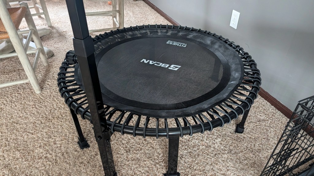 A close-up view of a black rebounder trampoline with a circular jumping surface and sturdy legs, positioned on carpeted flooring.