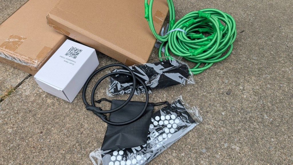 A collection of items on a concrete surface, including two cardboard boxes, a small white box with a QR code, several green cables, and black items wrapped in plastic.