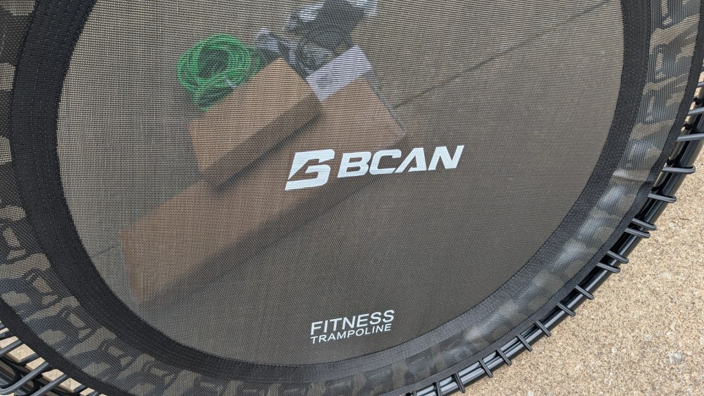 Close-up of a BCAN fitness trampoline featuring the brand name and 'FITNESS TRAMPOLINE' printed on the mat, with workout equipment visible in the background.