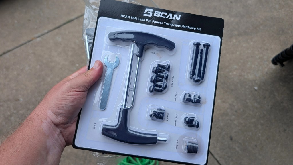 A person holding a BCAN Soft Land Pro Fitness Trampoline Hardware Kit, featuring various tools and hardware pieces, including a T-handle, wrench, and screws, packaged in a clear plastic display.