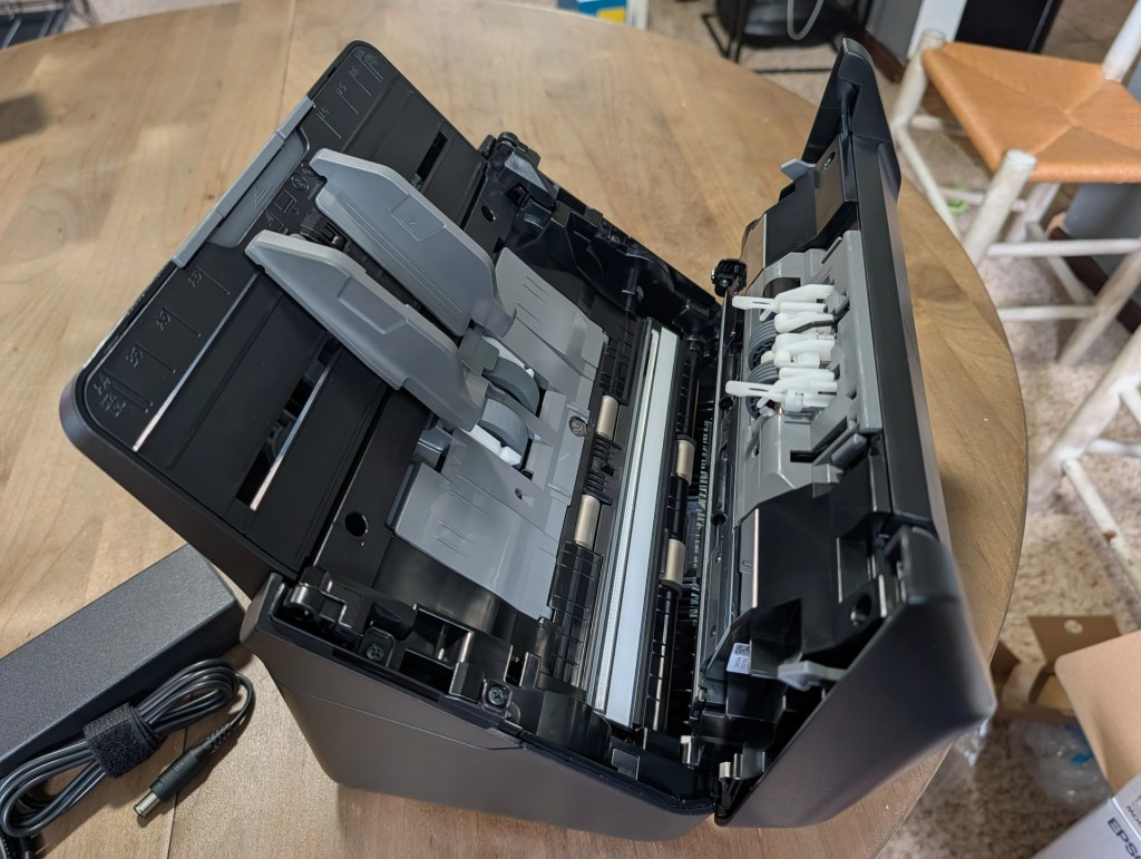 An open scanner showing its internal components on a wooden table, with a power supply cord nearby.