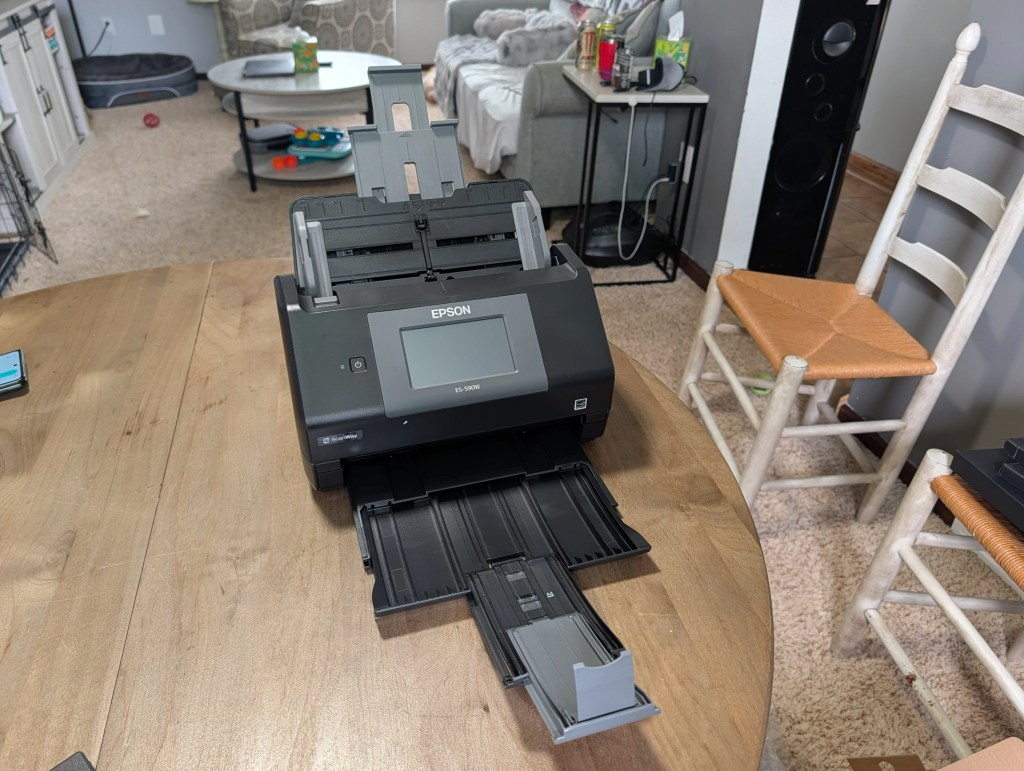 Epson document scanner on a wooden table, with a view of a living room in the background, including a couch and nearby furniture.