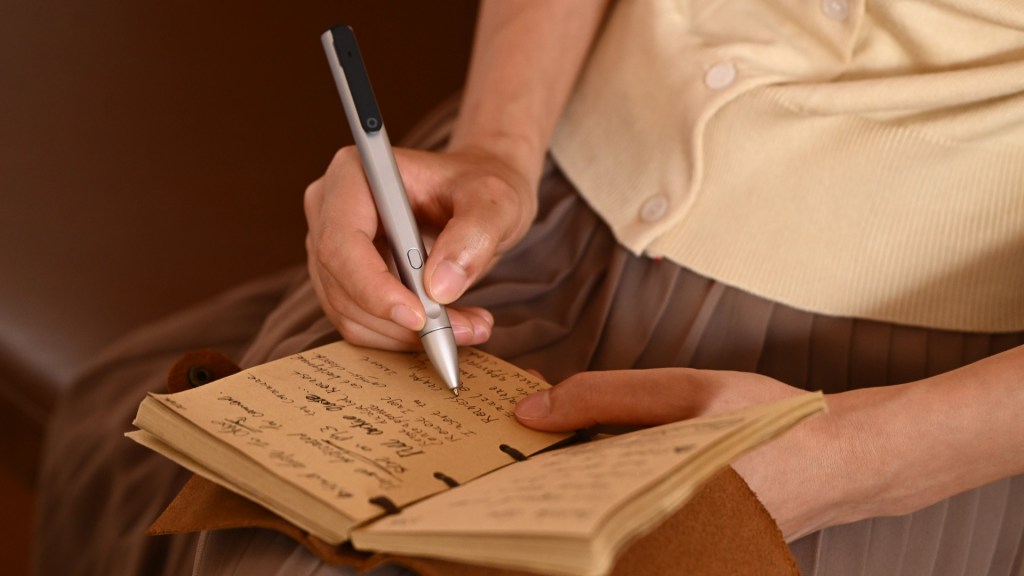A person writing in a notebook using a stylus, seated with a soft background. The notebook has handwritten notes and the individual is wearing a light sweater.