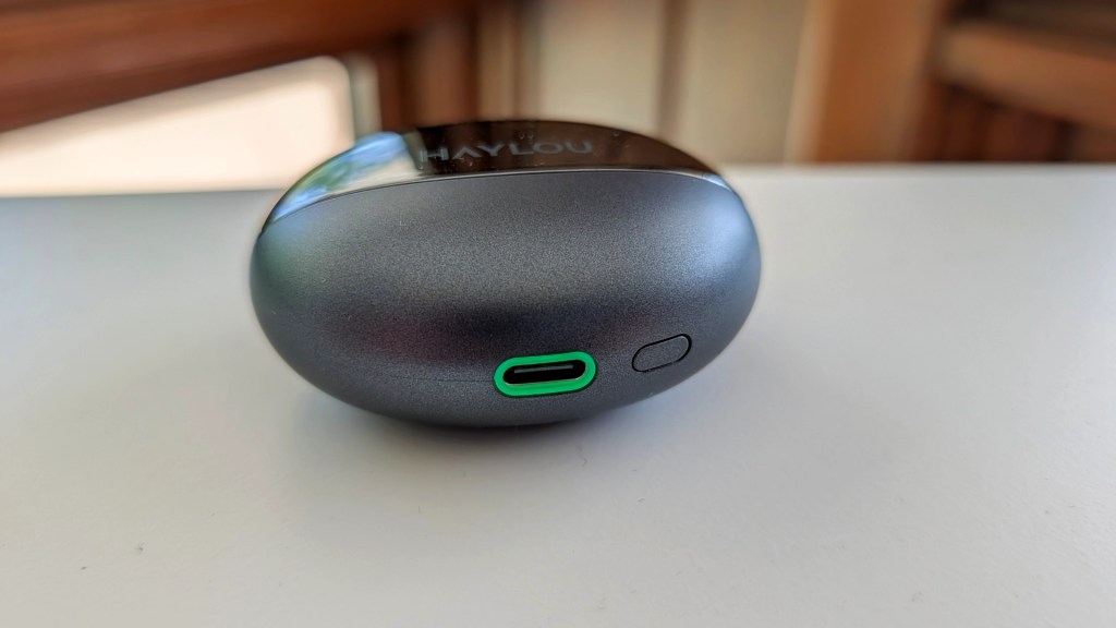 Close-up of a gray wireless earbud charging case showing a USB-C port and a button, placed on a white surface.