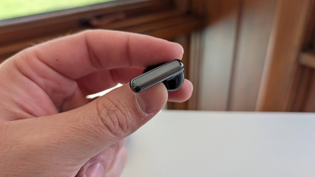 A hand holding a small black electronic device, possibly a wireless earbud, with a blurred background of wooden features.
