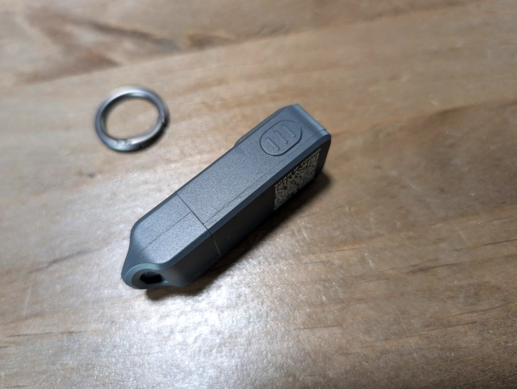 A gray portable device, likely a key fob or USB stick, next to a metal keyring on a wooden surface.