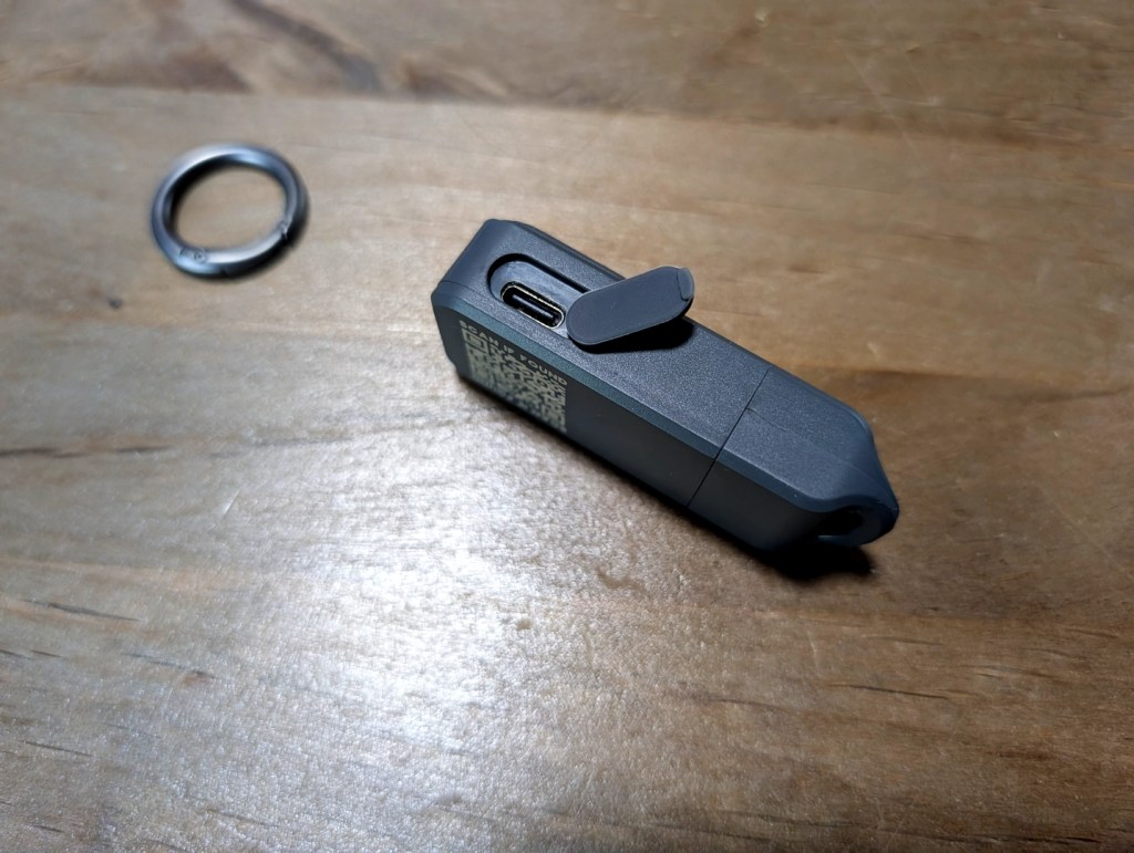 A close-up of a portable device with a USB-C port and a protective cover, placed on a wooden surface next to a metal keyring.