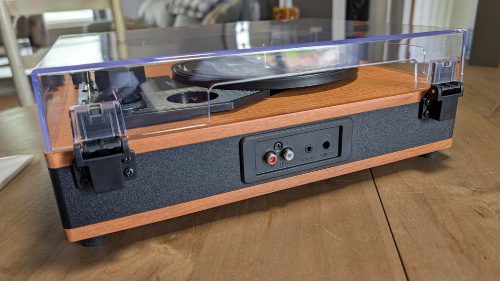 A side view of a vintage-style turntable with a wooden base and clear dust cover, placed on a wooden table. The back panel shows various audio input options.