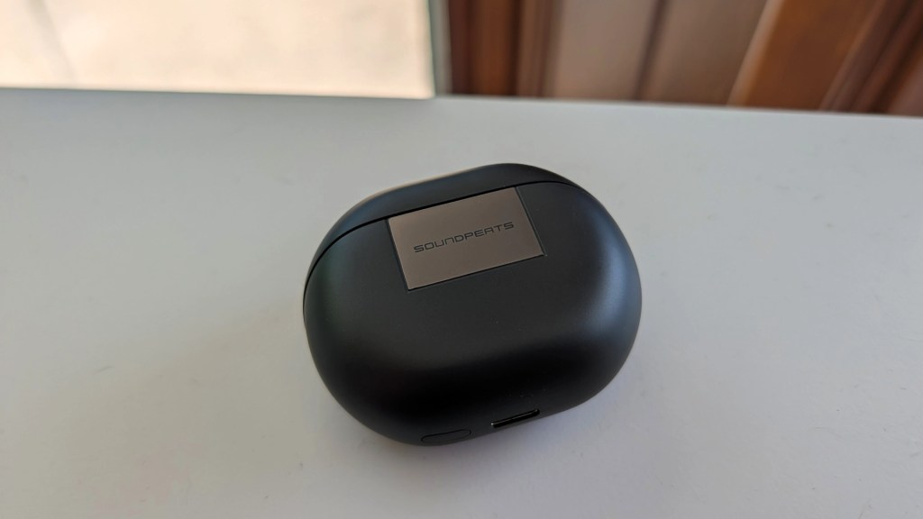 A black wireless earbud charging case with a metallic logo on top, resting on a white surface.