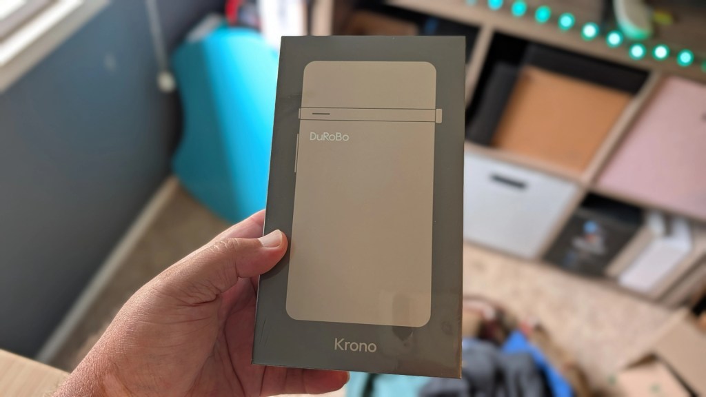 A person holding a sealed package of the DuRoBo Krono product, featuring a minimalist design and light color scheme.