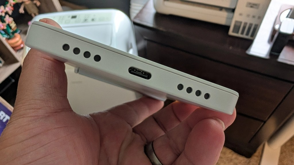 A person holding a white device featuring multiple speaker holes and a USB-C charging port, with a desk and decorative plants visible in the background.