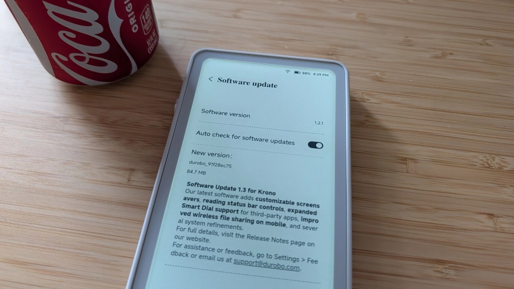 A smartphone displaying a software update screen with version details and options, next to a can of Coca-Cola on a wooden surface.