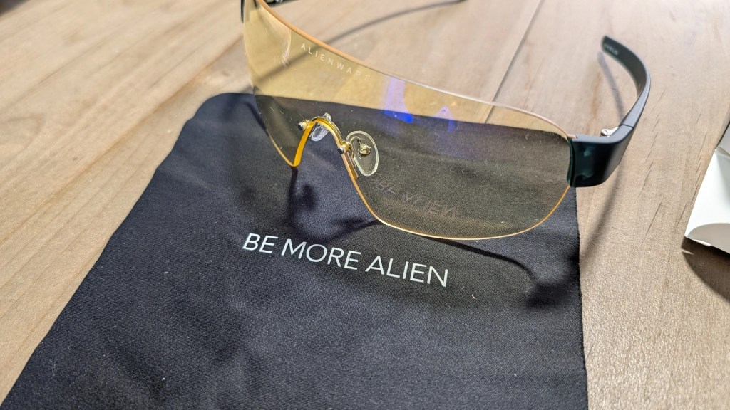 A pair of gaming glasses with yellow lenses placed on a black cloth displaying the phrase 'BE MORE ALIEN.'