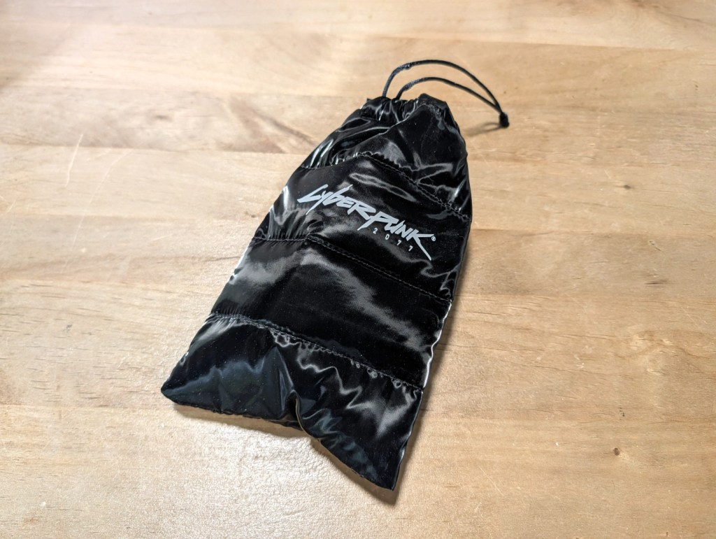 A black drawstring pouch with the 'Cyberpunk 2077' logo printed in white on its surface, resting on a wooden table.