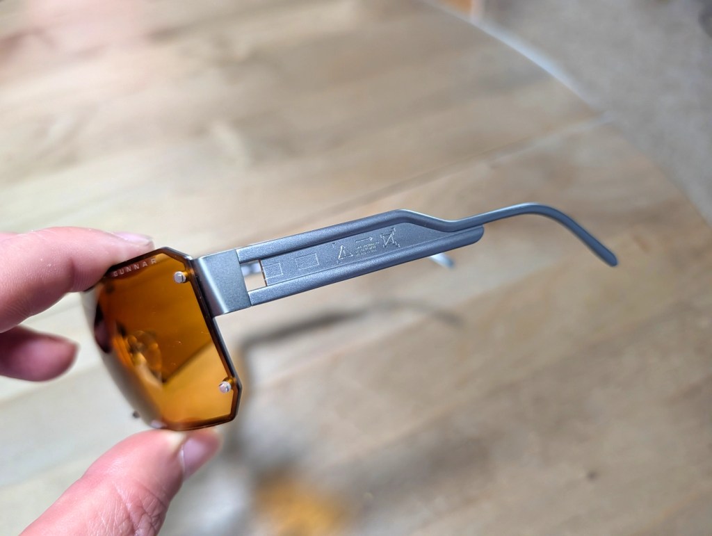 A person holding a pair of hexagonal amber sunglasses, showcasing the frame and side arm details.