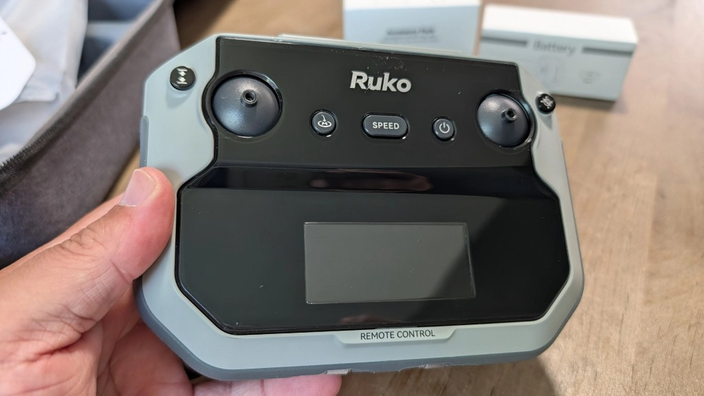 Close-up of a hand holding a Ruko remote control, featuring two joysticks, buttons for speed and mode, and a display screen.