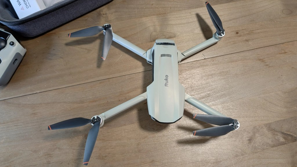 A compact drone with four propellers displayed on a wooden surface, next to a storage case.