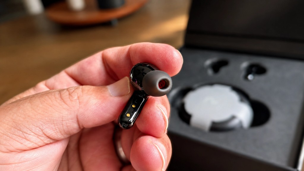 A person holding a black wireless earbud with a rubber tip, with a charging case visible in the background.
