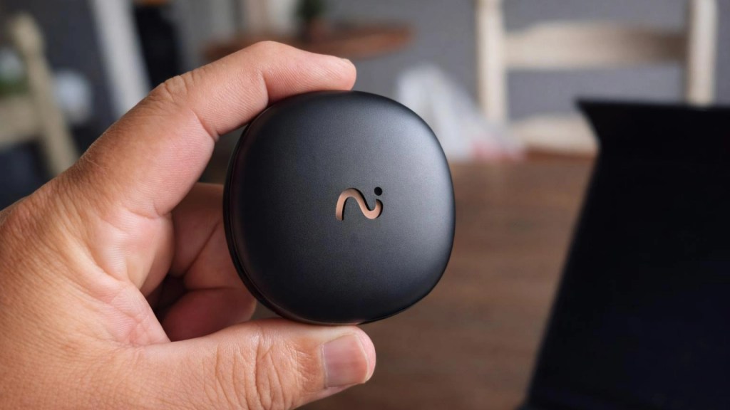 A close-up of a person's hand holding a small black electronic device with a logo on the front, set against a blurred background.