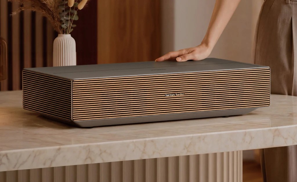 A hand resting on a stylish speaker with a grey finish and wooden accents, placed on a marble table, surrounded by decorative elements.