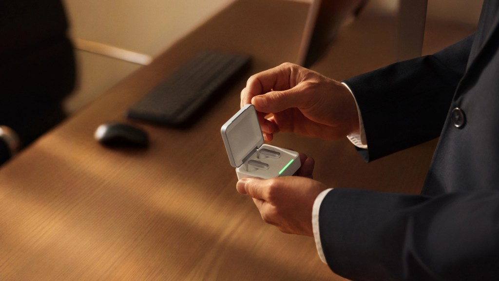 A person in a suit holding a wireless earbud case, partially opened, on a wooden desk with a computer keyboard in the background.